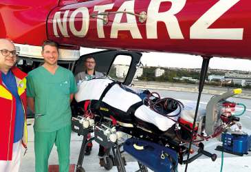 Notarztteam mit Patient auf Trage und ECMO-System vor Rettungshubschrauber auf dem Dachlandeplatz einer Klinik.