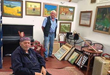 Zwei Männer in einer Kunstgalerie mit zahlreichen Gemälden – Gespräch über Malerei und kreative Werke.