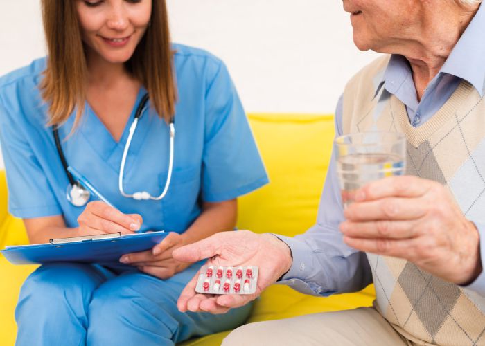 Pflegekraft dokumentiert Medikation, älterer Patient hält Tablettenblister und Glas Wasser zur Einnahme bereit